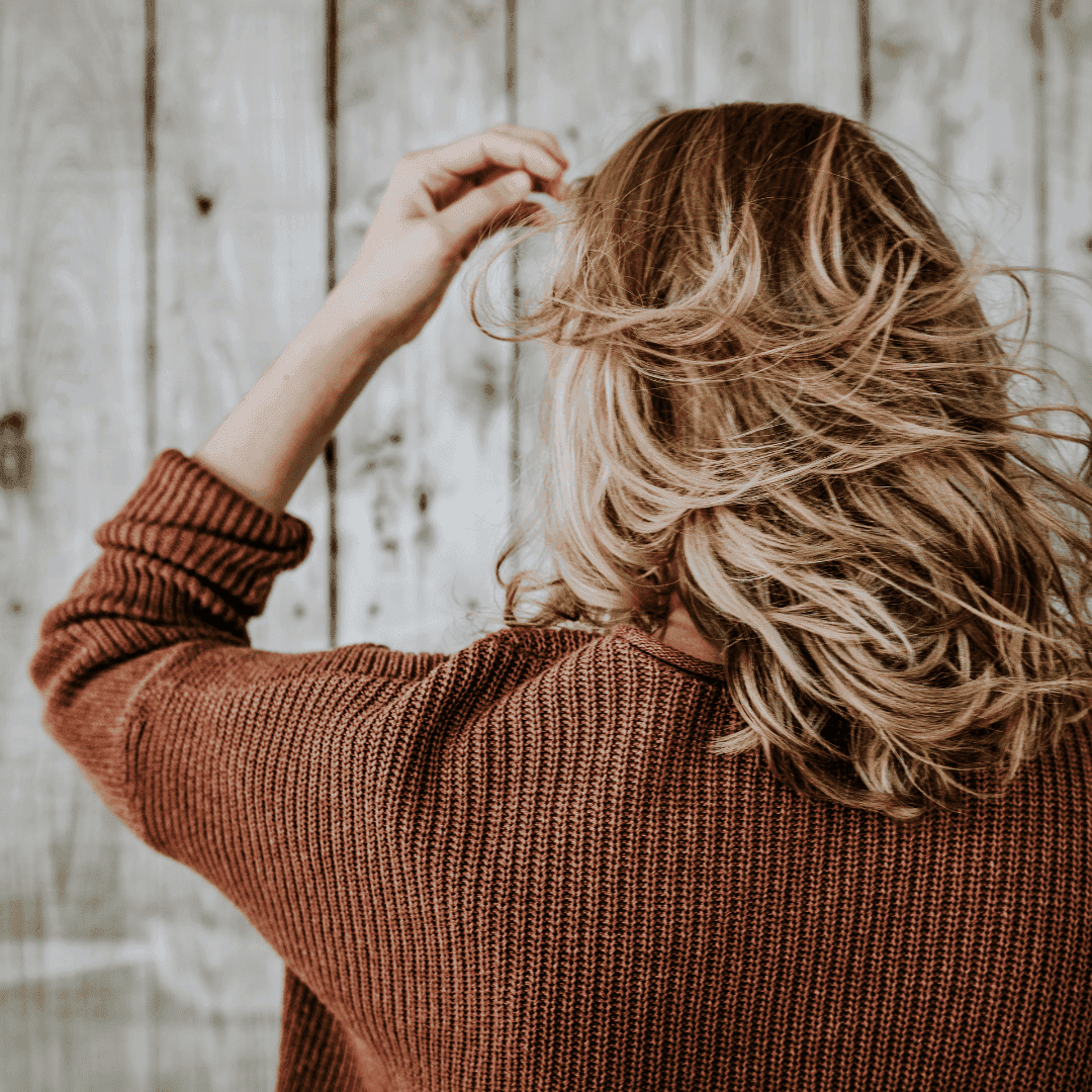 Woman with wavy hair in brown sweater facing wooden wall.