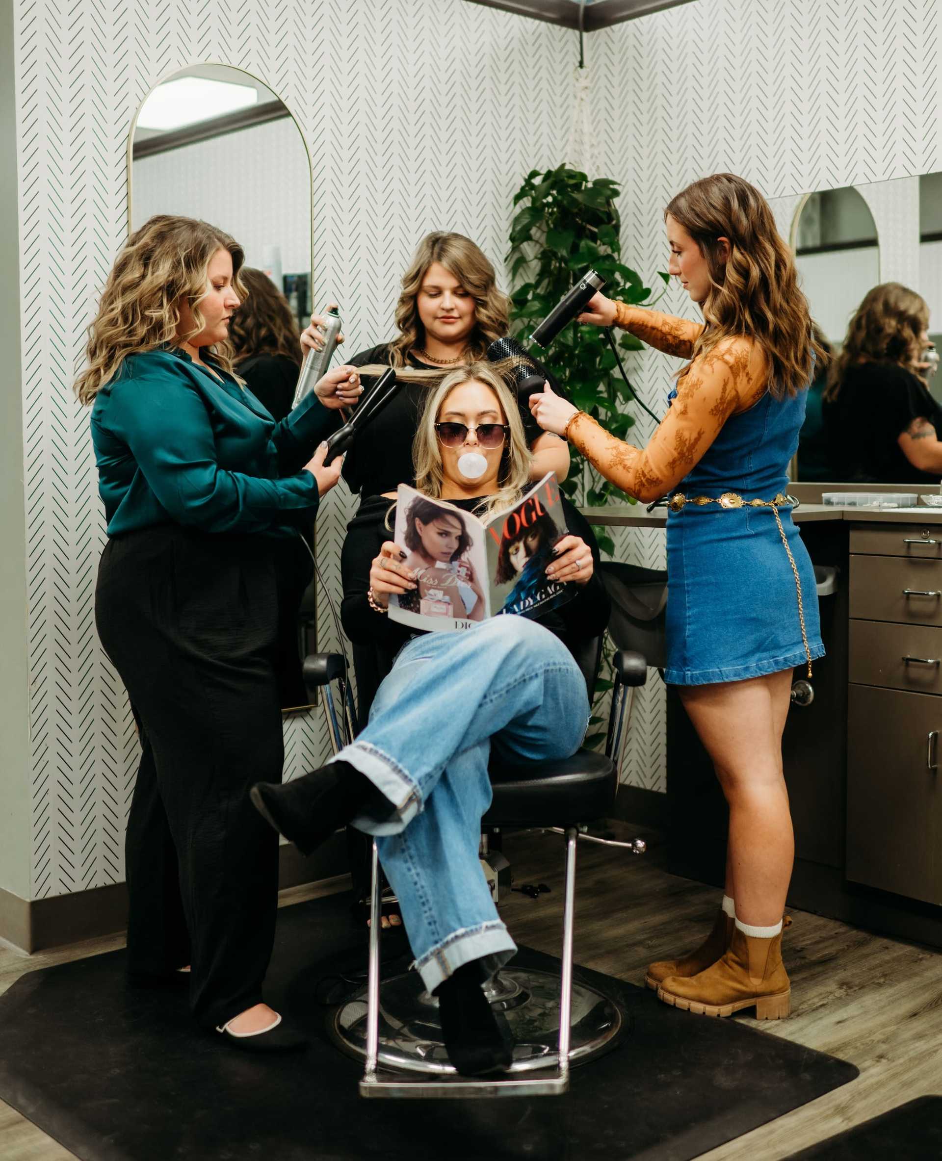 Woman sits in salon chair reading Vogue while getting hair styled by three stylists.