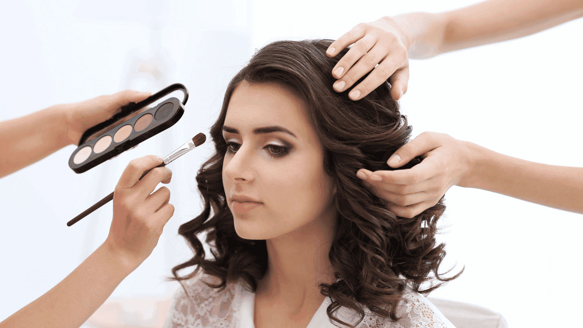 Makeup artist applying eye shadow to a woman with curling hair styled by another assistant.