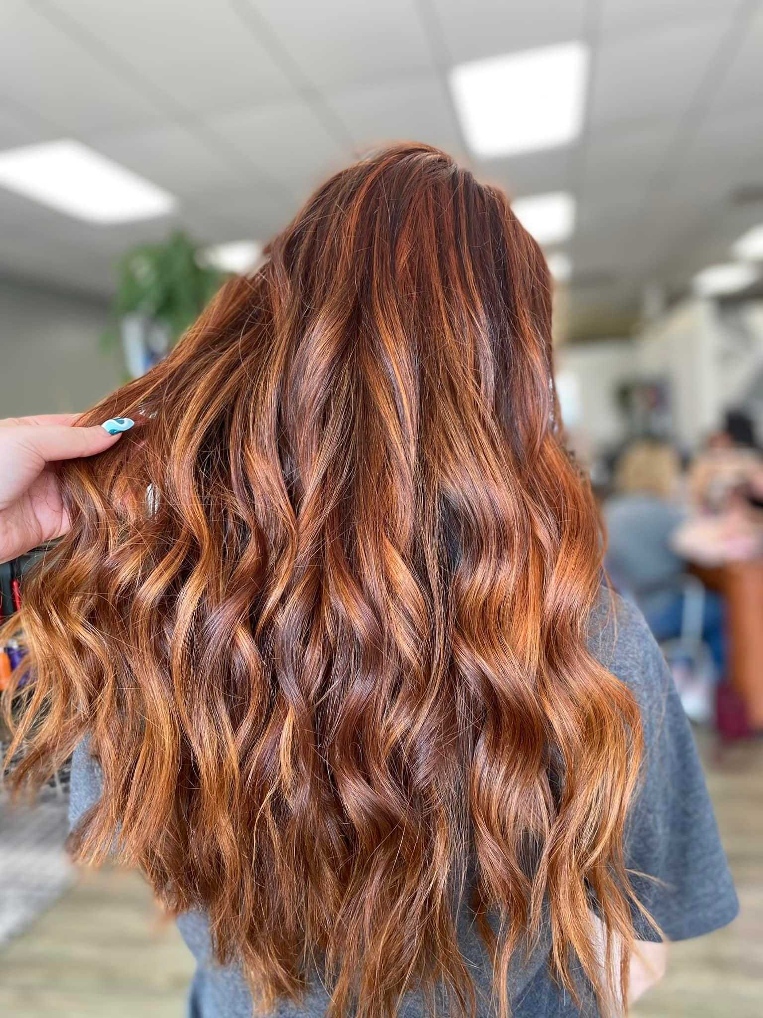 Close-up of a person with long, wavy red hair in a salon, being held by a hand with blue nails.