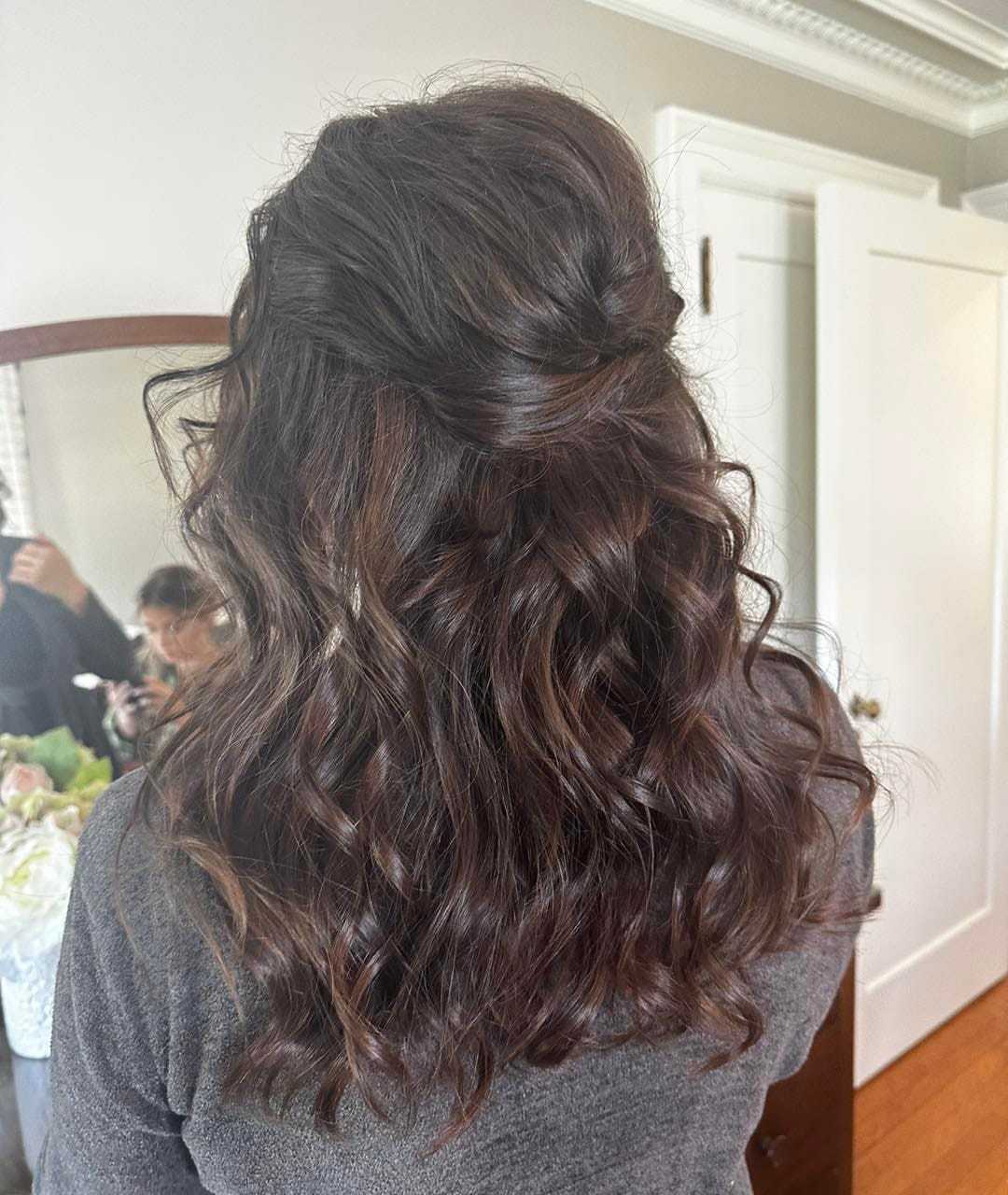 Woman with loose, wavy hairstyle in a gray top, in a room with white walls and a mirror.